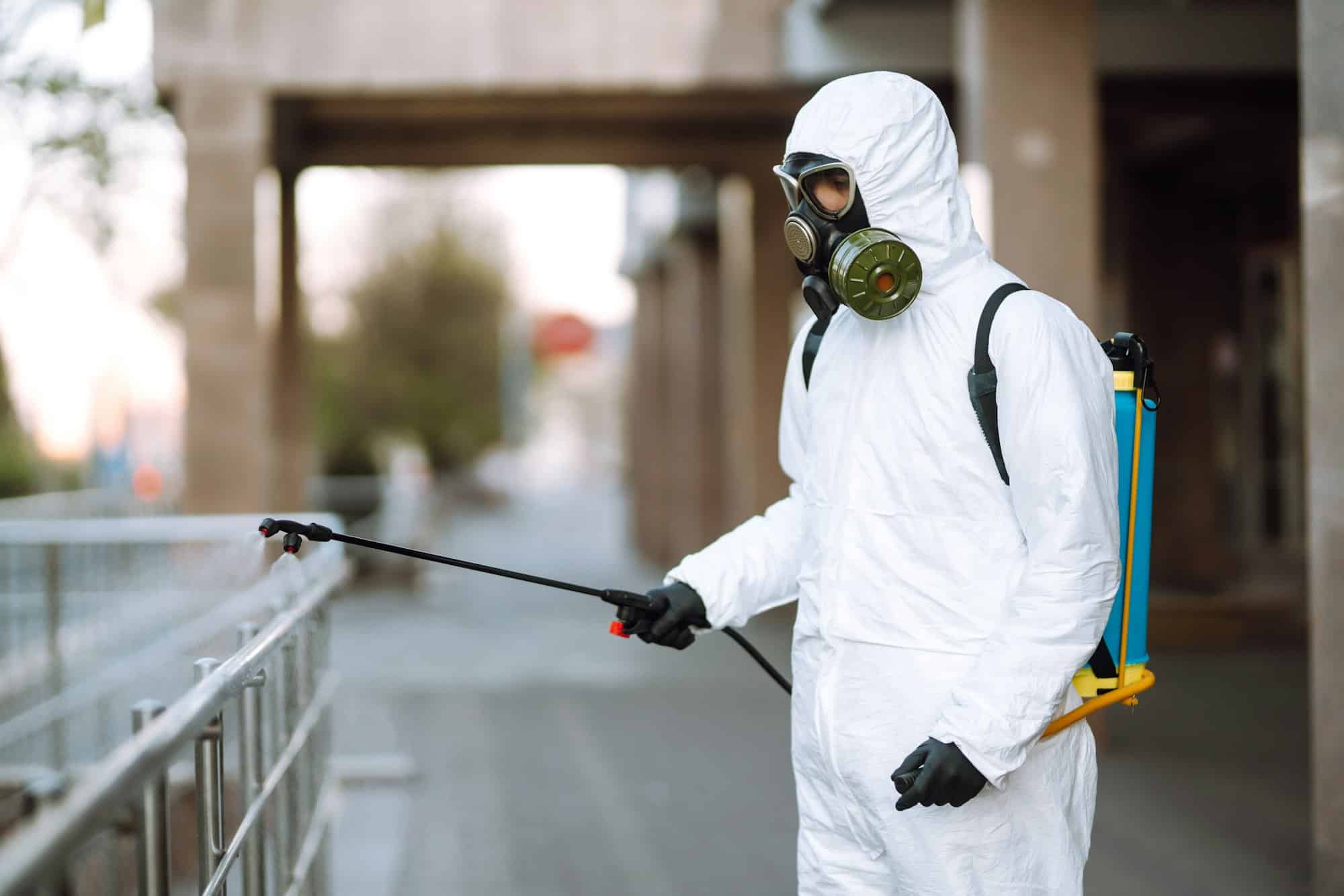 Man in protective suit and mask sprays disinfector onto the railing in the public place. Covid -19.